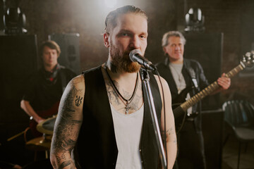Waist up portrait of bearded Caucasian man with tattoos on his body singing in microphone and looking at camera during performance with rock band