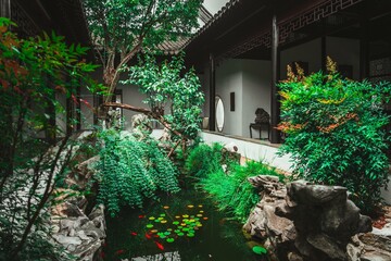 A lush, green garden with a small pond surrounded by rocks, trees, and traditional Chinese...