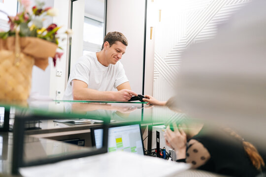 Remote view of male customer using credit card to pay on contactless NFC terminal at clinic counter. Handsome man making contactless payment with card at machine terminal at hotel reception.