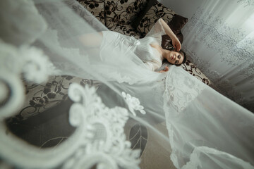 A woman in a white dress is laying on a bed with a white lace curtain behind her. The curtain is...