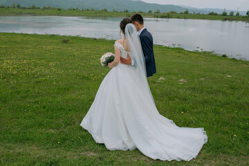 A bride and groom are walking together in a field near a body of water. The bride is holding a bouquet and the groom is wearing a suit. The scene is peaceful and romantic