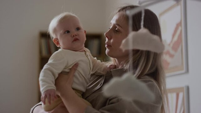Medium shot of young Caucasian woman holding baby in bedroom, kissing on cheeck and attracting attention to plush mobile, encouraging little child for exploration