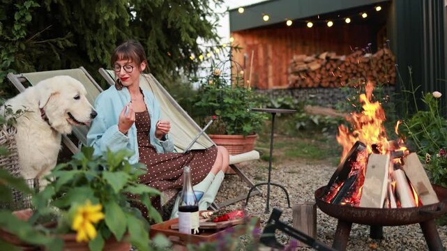 A woman enjoys a cozy evening watching a movie on her tablet, with her loyal dog by her side, as they sit by the warmth of a crackling fire in the serene garden setting