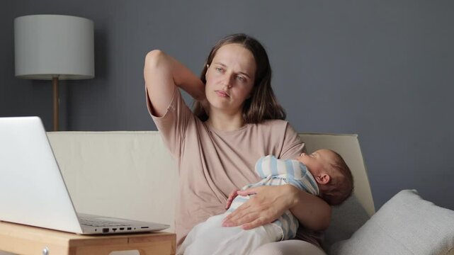 Exhausted mom sitting on couch with her baby using her laptop to work online while her toddler sleeping balancing remote work with family time massaging painful neck