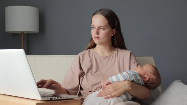 Smiling woman wearing beige shirt sitting on sofa with baby and rocking him female typing on notebook keyboard and looking after her r infant daughter while doing her remote job