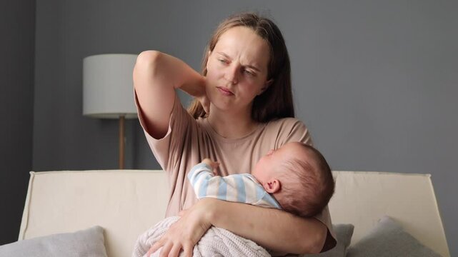 Sad upset Caucasian woman parent putting her infant son or daughter to sleep sitting on sofa lulling the baby to sleep while sitting in the cozy living room suffering painful neck grimacing from pain