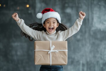 Obraz premium A cheerful child in a Santa hat eagerly raises a wrapped Christmas gift in excitement, embodying the joy and wonder of the holiday season in a festive setting.
