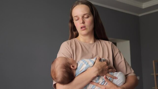 Tired exhausted Caucasian woman wearing casual clothing mommy rocking her newborn baby while standing in the living room holding infant son or daughter trying to put the child to bed