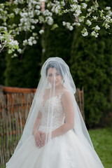 A woman in a wedding dress is standing in front of a tree with white flowers. She is wearing a veil and a veil cover