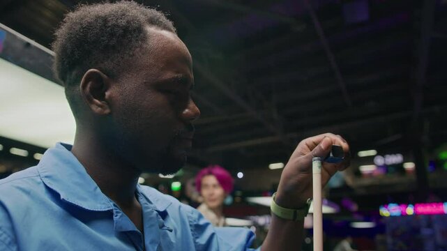 Chest up of young African American man calking his cue while playing billiards in bar at night