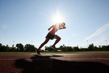 Against blinding sun, athlete in red launches into sprint, his form sharp and focused as he races toward his goal on open track. Concept of sport, tournament, motivation, workout, competition. Ad