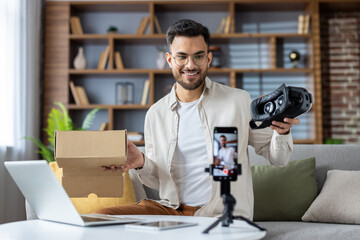 Young man creating unboxing video content with virtual reality headset and smartphone in modern...