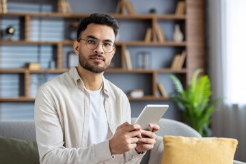 Confident young man holding tablet in modern living room with plants and shelves, wearing casual attire and glasses