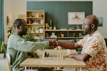 Side view shot of two young African American male chess players wearing casual clothes sitting at wooden table greeting each other with handshake