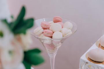 A glass bowl filled with pink and white macarons. The macarons are arranged in a way that they look like they are in a martini glass. The bowl is placed on a table next to a vase of flowers