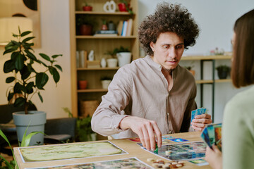 Young biracial man with curly hair sitting at table in living room and playing board game with unrecognizable girl friend