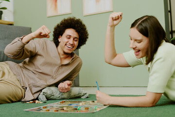 Cheerful young ethnically diverse couple having fun playing board game on floor in living room