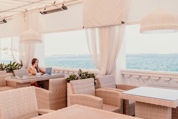 Woman coffee cafe laptop sea. Female freelancer using laptop on coffee break in modern cafe with sea view in the morning