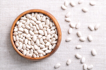 White beans in a wooden bowl on linen fabric. Dried small seeds, a variety of Phaseolus vulgaris, the common bean, a member of the legume family Fabaceae. Vegetarian staple food. Close up, from above.