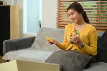 Happy young woman holding glass jar filled with coins and bank passbook sitting on sofa. Saving...