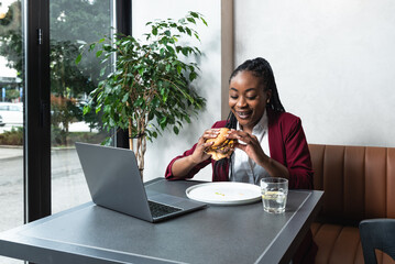 Young business woman working remotely from cafeteria during lunch break. Female office worker using laptop computer while she eating at restaurant to work or watching funny content on internet.
