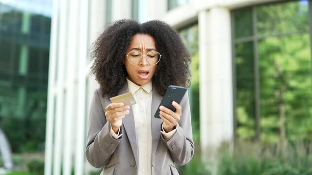 African American businesswoman holding a credit card looking at phone with stressed expression outside an office building. Woman has detected fraud or is worried about security of online transactions