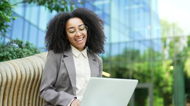 African american businesswoman smiling at laptop while sitting outdoors on bench near office business building, expressing excitement and joy. Concept of success, celebration, and good news. Close up