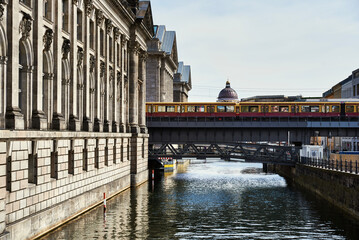 Naklejka premium Yellow metro in Berlin stopped over a bridge on the river Spree with the castle dome standing out. Historical old building by the canal with the subway standing by on a bridge. Berlin tourism concept
