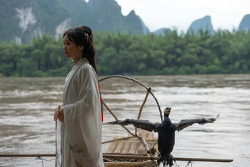 Hanfu girl smiles while seated on a bamboo raft, holding a round fan with cormorant birds beside her