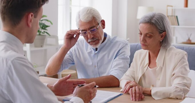 Senior couple at desk in office listening to young male realtor for property offer, sale to elderly spouses old customers reading contract on table, plan to take bank loan or medical health insurance 