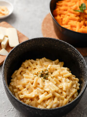 Two black bowls of macaroni and cheese on a gray surface. The front bowl contains creamy mac and cheese, garnished with herbs. In the background, a second bowl features a different cheese sauce