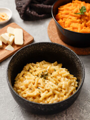 Two black bowls of macaroni and cheese on a gray surface. The front bowl contains creamy mac and cheese, garnished with herbs. In the background, a second bowl features a different cheese sauce