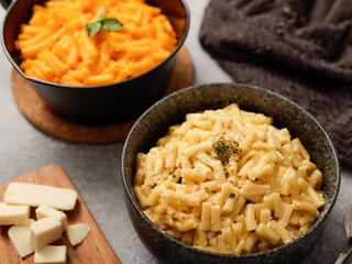 Two black bowls of macaroni and cheese on a gray surface. The front bowl contains creamy mac and cheese, garnished with herbs. In the background, a second bowl features a different cheese sauce