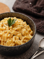 A close-up shot of a black bowl filled with creamy macaroni and cheese, garnished with rosemary. The bowl sits on a gray textured surface, with cheese slices on a wooden board in the background.