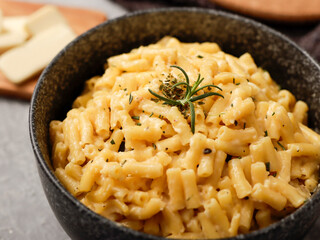 A close-up shot of a black bowl filled with creamy macaroni and cheese, garnished with rosemary. The bowl sits on a gray textured surface, with cheese slices on a wooden board in the background.