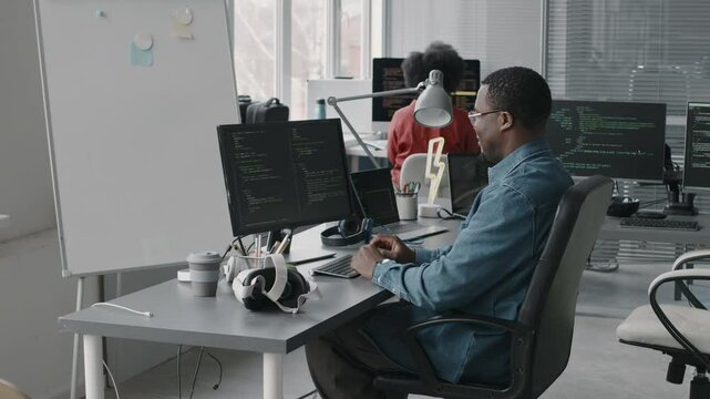 Side view medium shot of African American male software engineer typing html program code in computer while working in modern IT office with colleague