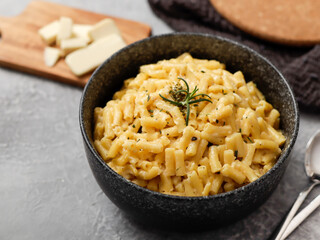 A close-up shot of a black bowl filled with creamy macaroni and cheese, garnished with rosemary. The bowl sits on a gray textured surface, with cheese slices on a wooden board in the background.