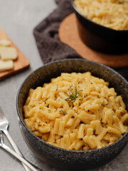 Two black bowls filled with creamy macaroni and cheese, garnished with rosemary, sit on a gray textured surface. Cheese slices on a wooden board are visible in the background a dark cloth.