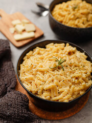 Two black bowls filled with creamy macaroni and cheese, garnished with rosemary, sit on a gray textured surface. Cheese slices on a wooden board are visible in the background a dark cloth.