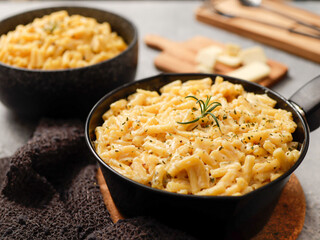 Two black bowls filled with creamy macaroni and cheese, garnished with rosemary, sit on a gray textured surface. Cheese slices on a wooden board are visible in the background a dark cloth.