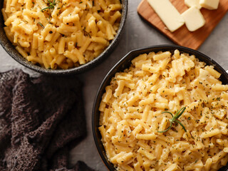 Two black bowls filled with creamy macaroni and cheese, garnished with rosemary, sit on a gray textured surface. Cheese slices on a wooden board are visible in the background a dark cloth.