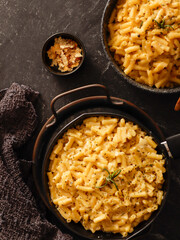 Two black bowls filled with creamy macaroni and cheese, garnished with rosemary, sit on a gray textured surface. Cheese slices on a wooden board are visible in the background a dark cloth.