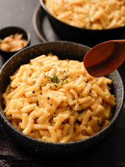 Two black bowls filled with creamy macaroni and cheese, garnished with rosemary, sit on a gray textured surface. Cheese slices on a wooden board are visible in the background a dark cloth.
