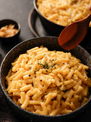 Two black bowls filled with creamy macaroni and cheese, garnished with rosemary, sit on a gray textured surface. Cheese slices on a wooden board are visible in the background a dark cloth.