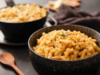Two black bowls filled with creamy macaroni and cheese, garnished with rosemary, sit on a gray textured surface. Cheese slices on a wooden board are visible in the background a dark cloth.