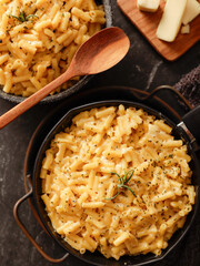 Two black bowls filled with creamy macaroni and cheese, garnished with rosemary, sit on a gray textured surface. Cheese slices on a wooden board are visible in the background a dark cloth.
