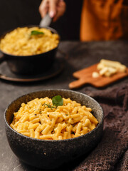 A black bowl filled with creamy macaroni and cheese, garnished with fresh basil leaves, sits on a dark surface. A blurred hand in the background is holding another bowl of the same dish.