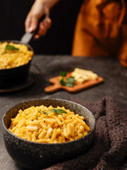 A black bowl filled with creamy macaroni and cheese, garnished with fresh basil leaves, sits on a dark surface. A blurred hand in the background is holding another bowl of the same dish.