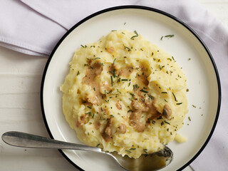 A plate of creamy mashed potatoes topped with a rich, savory gravy and garnished with fresh herbs. The dish is served on a white plate, with a spoon placed on the side, ready to be enjoyed