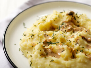 A plate of creamy mashed potatoes topped with a rich, savory gravy and garnished with fresh herbs. The dish is served on a white plate, with a spoon placed on the side, ready to be enjoyed